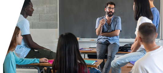 Image of male teacher at front of classroom, leaning against desk talking with students