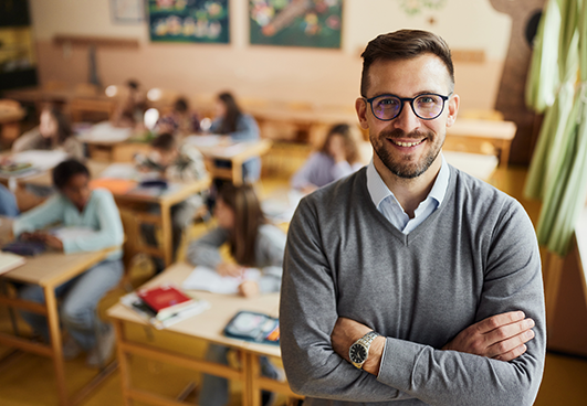Image of smiling man standing in front of classroom, facing the camera with arms crossed